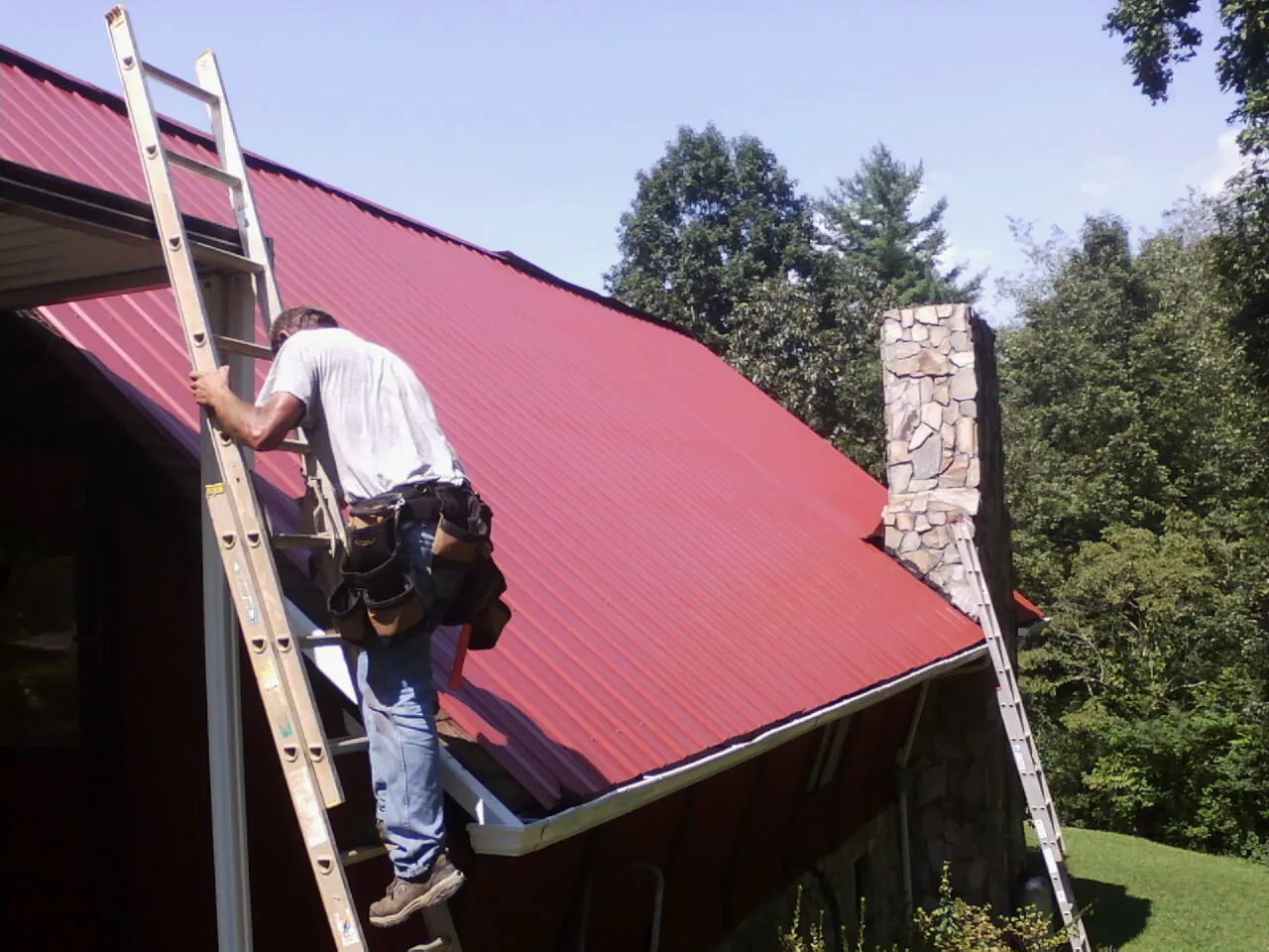 Roofer on a ladder beside a red metal roof and stone chimney — Soffit & Fascia Repair by Roofing Brothers in Mission Viejo